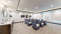 Located at 4351 Little Egret Road in Kissimmee, Florida, this sophisticated single-floor living room functions as a refined screening and gathering space. The room is thoughtfully designed with a coffered tray ceiling accented by dark wood trim and recessed lighting, creating a layered, upscale ambiance throughout.

The seating arrangement features rows of plush blue-and-white armchairs with wooden accents and integrated tablet arms, oriented toward a large flat-screen television mounted above a warm-toned wood credenza. The credenza provides generous storage and display space, complemented by decorative greenery and subtle accessories that enhance the room's polished aesthetic.

A patterned, neutral-toned carpet with a delicate botanical motif covers the floor, adding texture and warmth underfoot. Ambient teal accent lighting along one wall introduces a modern, cinematic quality to the space. Cinema-themed wall art adds thematic character, reinforcing the room's entertainment focus.

Toward the rear of the room, a built-in wet bar or service counter with dark wood cabinetry, a white countertop, and a sink is flanked by elegant wall sconces and an arched mirror, offering a functional yet stylish refreshment area. A wide, dark-framed entry door with keypad access provides secure, controlled entry. The overall design balances comfort, functionality, and contemporary elegance.