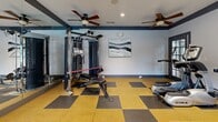 Located at 2090 Columns Dr in Cumming, Georgia, this fitness center offers a well-equipped exercise space designed to support a well-rounded workout routine. The room features a distinctive checkered rubber flooring in yellow and dark gray tones, providing both durability and a polished aesthetic. Two ceiling fans with integrated lighting ensure consistent air circulation throughout the space, complementing the recessed lighting overhead.

The fitness center is outfitted with a multi-functional cable machine and adjustable bench for strength training, a dumbbell rack with a range of free weights, and cardio equipment including a treadmill, elliptical, and a rowing machine. A large wall-mounted mirror on one side of the room enhances the sense of space and allows users to monitor their form during exercise. A wall-mounted flat-screen television adds entertainment options during workouts.

Natural light fills the room through multiple large windows with dark-framed trim, and a glass-panel door provides direct access to an outdoor pool area, offering pleasant views of the surrounding amenity space. A second entrance door allows convenient access from the exterior. The crisp white walls are accented by a bold dark navy border trim, giving the space a clean and modern appearance. Posted fitness center rules reflect the organized and community-oriented nature of this facility.