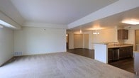 This single-floor residence showcases an open-concept layout that connects the main living area, dining space, and kitchen in one cohesive flow. The living room is finished with neutral-toned wall-to-wall carpeting, creating a soft and comfortable foundation, while the adjoining dining and entry areas transition to darker hard-surface flooring, providing a clear visual distinction between zones.

The living space features smooth white ceilings with a subtle tray ceiling detail, adding a touch of architectural dimension. Neutral walls throughout contribute to a bright, airy atmosphere. A small chandelier anchors the dining area, adding a classic decorative element, while a flush ceiling fixture provides additional lighting near the entryway.

The kitchen area is partially visible and features a raised countertop with stone-look surfaces, functioning as a convenient breakfast bar or pass-through that connects the kitchen to the dining space. A closet near the main entry offers practical storage. The overall palette of whites and neutrals lends the space a clean, versatile aesthetic well-suited for a variety of interior styles.
