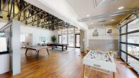 Located at 170 Graphics Dr in Madison, Alabama, this single-floor clubhouse at Livano Madison is a thoughtfully designed community amenity space that blends industrial character with modern comfort. The space is defined by its dramatic, soaring ceilings with exposed dark steel roof trusses — a striking architectural feature that adds both visual depth and an open, airy atmosphere throughout. Warm hardwood flooring runs continuously across the entire floor, unifying the distinct functional zones within the space.

The main game and recreation area is generously proportioned, accommodating a billiards table, a ping pong table, and a foosball table, making it an ideal gathering spot for social activity. Large floor-to-ceiling windows and glass-paneled roll-up garage doors flood the space with abundant natural light while providing seamless access to the outdoor patio area beyond.

A separate kitchen and dining area features a long, contemporary island with bar seating, built-in shelving, a sink, and a wall-mounted television. Full-panel glass garage doors in this zone further enhance the indoor-outdoor connection and maximize natural light.

The lounge area near the Livano leasing reception desk is elegantly appointed with tufted leather ottomans, upholstered sofas, and refined dining furniture beneath the dramatic truss ceiling, creating a welcoming and sophisticated atmosphere for residents to relax and connect.