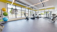 Located at 545–547 New Park Ave in West Hartford, CT, this fitness center offers a well-equipped, single-floor exercise facility designed with both functionality and an energizing aesthetic in mind. The space is characterized by its expansive floor-to-ceiling glass curtain walls that wrap around a significant portion of the perimeter, flooding the interior with abundant natural light and providing open views of the surrounding streetscape and greenery outside.

The facility's design features exposed industrial-style ductwork running along the white ceiling, complemented by bold yellow accent walls that add a dynamic, motivating energy to the environment. Durable speckled rubber flooring covers the entire space, providing a resilient, safety-conscious surface suited for a wide range of physical activities.

The cardio zone, positioned prominently along the windowed walls, is outfitted with treadmills, ellipticals, stationary bikes, and a rowing machine. A dedicated strength training area houses cable and weight machines, a multi-functional cable station, and a dumbbell rack. The open floor plan allows for ample spacing between equipment, creating a comfortable and organized workout environment. Entry is through a glass-paneled door, and the layout accommodates ease of movement throughout the entire facility.