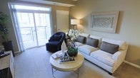 This single-floor living space showcases a thoughtfully designed interior with a neutral color palette of warm beige and taupe tones throughout. The living area serves as the focal point, anchored by a generously sized light-colored sectional sofa accented with charcoal gray throw pillows, complemented by a sleek, round coffee table in a champagne finish. A stylish black accent chair is positioned near the sliding glass door, which floods the room with natural light and provides direct access to a private balcony with views of the surrounding area. The balcony extends the living space outdoors, offering an inviting spot for relaxation. Wall-to-wall carpeting in a soft neutral tone adds warmth underfoot. A tall floor lamp with a clean white shade provides ambient lighting, while a framed decorative wall panel adds a refined artistic touch. Fresh orchids and carefully curated decorative accessories on the coffee table contribute to the polished, hotel-inspired aesthetic. A doorway visible in the background hints at additional rooms beyond, rounding out this well-organized and elegantly presented space.
