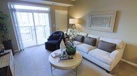 This single-floor living space showcases a thoughtfully designed interior with a neutral color palette of warm beige and taupe tones throughout. The living area serves as the focal point, anchored by a generously sized light-colored sectional sofa accented with charcoal gray throw pillows, complemented by a sleek, round coffee table in a champagne finish. A stylish black accent chair is positioned near the sliding glass door, which floods the room with natural light and provides direct access to a private balcony with views of the surrounding area. The balcony extends the living space outdoors, offering an inviting spot for relaxation. Wall-to-wall carpeting in a soft neutral tone adds warmth underfoot. A tall floor lamp with a clean white shade provides ambient lighting, while a framed decorative wall panel adds a refined artistic touch. Fresh orchids and carefully curated decorative accessories on the coffee table contribute to the polished, hotel-inspired aesthetic. A doorway visible in the background hints at additional rooms beyond, rounding out this well-organized and elegantly presented space.