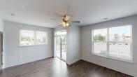 This single-floor residence showcases a clean, contemporary design with dark hardwood-style flooring running throughout. The living area is bright and open, benefiting from multiple windows with white blinds that allow generous natural light to filter in. A ceiling fan with a light fixture provides both comfort and illumination, while sliding glass doors lead directly to a private balcony with outdoor views, extending the functional living space.

The kitchen is well-appointed with dark espresso cabinetry reaching toward the ceiling, granite countertops, and a full suite of stainless steel appliances, including a refrigerator, range, microwave, and dishwasher. Flush-mount lighting illuminates the kitchen and entry corridor, which leads to a neutral-toned front door.

The bathroom features a granite-topped vanity with an undermount sink and dark cabinetry, paired with wood-look flooring. A grab bar is installed near the toilet, and large tile panels line the walls. A doorway from the bathroom provides direct access to a walk-in closet with built-in shelving and hanging rods. Additional rooms maintain the consistent light gray wall palette and dark flooring, contributing to a cohesive, modern aesthetic throughout the space.