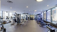 Located in Cleveland, OH, this fitness center spans a generously sized single-floor layout designed to accommodate a full range of workout needs. The space is divided into distinct functional zones, beginning with a bright, welcoming reception and lounge area featuring light hardwood flooring, plush seating, a reception desk, and floor-to-ceiling windows that frame striking views of the Cleveland skyline. Lush greenery and a relaxed seating arrangement give this entry area a comfortable, professional atmosphere.

Beyond the reception, the main workout floor opens into a spacious and well-lit gym area. Large windows line the perimeter, flooding the space with natural light and providing panoramic city views—a standout feature that elevates the exercise experience. The gym is outfitted with an extensive selection of strength-training machines, cable systems, free weights, a power rack, and a punching bag station for combat-style training. Durable rubber flooring and wall-length mirrors enhance both safety and the sense of openness throughout.

A dedicated cardio zone features a row of modern treadmills, ellipticals, and stationary bikes positioned directly in front of expansive windows overlooking the city. A separate area near the entrance accommodates indoor cycling bikes. Recessed and panel lighting throughout ensures consistent illumination across all workout zones, making this fitness center a comprehensive and inviting facility.