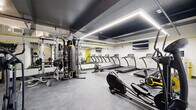 This fitness center offers a thoughtfully designed workout space across a single floor, providing ample room for a full range of exercise activities. The interior showcases a modern aesthetic defined by crisp white walls, a dramatic dark ceiling, and striking yellow accent stripes that run along the perimeter — creating a high-energy, motivating atmosphere.

The cardio zone is well-stocked with multiple treadmills, elliptical trainers, and a stationary spin bike, all positioned along mirrored walls that amplify the sense of space. A wall-mounted flat-screen TV enhances the cardio experience. The strength training area features a multi-station cable machine, a punching bag, kettlebells, medicine balls, and a rowing machine, catering to a diverse range of training styles.

Recessed linear LED lighting runs across the ceiling, providing bright, even illumination throughout the space. The floor is finished with durable rubber gym flooring, offering both safety and comfort. Large mirrors line the walls, reflecting light and giving the space an open, expansive feel. A glass-paneled entry door and a bold yellow accent door contribute to the facility's contemporary design. The overall ambiance is professional, clean, and purpose-built for an effective fitness experience.