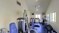This fitness center is thoughtfully designed to provide a comprehensive workout environment within a compact, single-floor layout. The space features vaulted ceilings that lend an open, airy feel, complemented by a ceiling fan that promotes airflow and comfort during workouts. Natural light filters in through a window at the far end of the room, creating a welcoming atmosphere.

The gym is outfitted with a solid selection of equipment to accommodate a range of fitness goals. Cardio options include multiple treadmills and an elliptical machine, all positioned to take advantage of the natural light and mounted flat-screen televisions for entertainment during workouts. For strength training, the space includes a multi-station cable and weight machine, ideal for targeting various muscle groups.

The dark rubber flooring provides both safety and durability underfoot, while the light-toned walls keep the space feeling clean and energized. Wall-mounted televisions are positioned for easy viewing from multiple exercise stations. A wall clock and posted instructional signage add functional detail to the space. Overall, this fitness center offers a practical, well-organized environment with all the essential amenities needed for an effective daily workout routine.