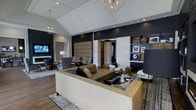 This single-floor space showcases a thoughtfully designed interior that balances bold aesthetics with functional elegance. The main living area commands immediate attention with its dramatic dark navy accent wall, which serves as a striking backdrop against lighter surrounding tones. A tray ceiling with recessed lighting and geometric gold pendant chandeliers adds architectural depth and a sense of elevated verticality to the room.

The seating area is anchored by a generously sized cream leather sofa and complementary upholstered furnishings in neutral and plaid patterns, creating distinct yet cohesive conversation zones. Rich hardwood flooring runs throughout, grounding the space with warmth and continuity. Large upholstered wall panels introduce a textural element near the central corridor, adding a boutique-hotel quality to the design.

A built-in shelving unit along one wall displays curated artwork and decorative objects, reinforcing the space's gallery-like sensibility. Recessed lighting combined with stylish floor and table lamps ensures layered illumination throughout. The overall ambiance is one of modern luxury — polished, intentional, and designed to impress.