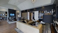 This single-floor space showcases a thoughtfully designed interior that balances bold aesthetics with functional elegance. The main living area commands immediate attention with its dramatic dark navy accent wall, which serves as a striking backdrop against lighter surrounding tones. A tray ceiling with recessed lighting and geometric gold pendant chandeliers adds architectural depth and a sense of elevated verticality to the room.

The seating area is anchored by a generously sized cream leather sofa and complementary upholstered furnishings in neutral and plaid patterns, creating distinct yet cohesive conversation zones. Rich hardwood flooring runs throughout, grounding the space with warmth and continuity. Large upholstered wall panels introduce a textural element near the central corridor, adding a boutique-hotel quality to the design.

A built-in shelving unit along one wall displays curated artwork and decorative objects, reinforcing the space's gallery-like sensibility. Recessed lighting combined with stylish floor and table lamps ensures layered illumination throughout. The overall ambiance is one of modern luxury — polished, intentional, and designed to impress.