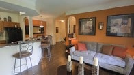 This single-floor residence showcases a thoughtfully designed open-concept layout that effortlessly connects the living, dining, and kitchen areas into one cohesive and inviting space. The interior is defined by rich, warm tones — most notably the bold terracotta-orange accent walls that lend a sense of energy and warmth throughout. Dark hardwood flooring runs continuously across the main living areas, providing a striking contrast against the white crown molding and architectural details that frame the ceilings and doorways. The living area features a comfortable upholstered sofa accented with coral throw pillows, complemented by a stylish coffee table adorned with decorative candle holders. An elegant arched doorway serves as a focal architectural detail, adding a classic touch to the open floor plan. Adjacent to the living space, the dining area includes a bar-height counter with modern stools, ideal for casual dining. The kitchen, visible from the main living space, is outfitted with sleek cabinetry and pendant lighting that adds a refined finishing touch. Wall art and decorative accents throughout contribute to the space's warm, curated aesthetic. The overall ambiance is one of comfort and style, making it a functional yet visually appealing home.