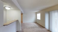 This single-floor space presents a clean, functional layout centered around an open living area. The room is finished with wall-to-wall carpet in a warm beige tone, complementing the neutral cream-colored walls throughout. A textured ceiling adds subtle visual interest overhead.

Natural light enters through a window on the far wall, brightening the space and contributing to an airy feel. The open layout offers flexibility for various furniture arrangements, making it adaptable to different living needs.

A partial wall and countertop overhang on the left side of the living area suggests an adjacent kitchen space, providing a natural visual separation between the two areas while maintaining an open feel. The countertop appears to feature a light-toned surface.

Visible in the background is a doorway leading to an additional room, fitted with light wood-toned bi-fold doors, likely a closet, hinting at practical storage solutions within the unit. Electrical outlets are positioned along the baseboards at convenient intervals.

Overall, the space maintains a simple, understated aesthetic with neutral finishes throughout, offering a blank canvas that is easy to personalize and furnish.
