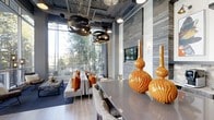 Located at 2610 Erwin Road in Durham, North Carolina, this striking single-floor space makes an immediate impression with its dramatic, loft-inspired design. Soaring ceilings draw the eye upward to an ornate laser-cut ceiling panel and an industrial-style exposed framework, creating a layered architectural experience that blends raw and refined elements seamlessly.

Floor-to-ceiling windows flood the interior with abundant natural light, framing lush outdoor greenery and fostering a strong connection between the inside and the surrounding landscape. The open-concept layout flows effortlessly from a dining area to a lounge space, anchored by a sleek, elongated dining table surrounded by woven upholstered chairs.

A statement wall clad in weathered wood planking adds warmth and texture, complemented by a wall-mounted flat-screen television and elegant sconce lighting. Pendant lights with a sculptural, globe-like metallic finish hang above the dining area, adding a bold focal point overhead.

The lounge area features plush neutral sofas accented with vibrant orange throw pillows, bringing energy and warmth to the cool grey palette. Lustrous gold ceramic vases serve as striking decorative centerpieces, reinforcing the space's commitment to curated, high-impact design. Light wood-tone flooring ties the entire aesthetic together with understated elegance.