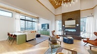 Located at 135 Ballentrae Ct in Chapel Hill, NC, this thoughtfully designed single-floor space blends modern farmhouse aesthetics with refined, contemporary details throughout. The open-concept layout connects the living, dining, and kitchen areas seamlessly, creating a fluid and welcoming environment filled with abundant natural light from expansive black-framed windows.

The living room is anchored by soaring vaulted ceilings clad in warm wood paneling and a dramatic floor-to-ceiling tiled fireplace, complemented by rich light hardwood flooring. Sage green cabinetry provides stylish storage and serves as a cohesive design thread throughout the space. A statement globe chandelier adds a refined focal point overhead.

The kitchen features warm natural wood cabinetry, striking marble-look wall tile that rises to the ceiling, sleek black appliances, and a distinctive hexagonal mosaic tile floor that adds artistic character. High clerestory windows flood the kitchen with natural light from above.

The dining area accommodates generous gatherings around a large communal table beneath a wood-paneled ceiling and modern linear chandelier, with built-in banquette seating along the walls.

A cozy lounge nook offers built-in green bench seating, a floor-to-ceiling bookshelf, tile flooring, and an oversized woven pendant light, creating an intimate retreat ideal for relaxing or reading. The entry area features light hardwood floors, curated artwork, and a welcoming, airy ambiance throughout.