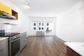Located at 104 S 11th St in Boise, Idaho, this single-floor unit presents a contemporary design with a well-considered layout. The kitchen is a standout feature, outfitted with bold yellow upper cabinetry accented by warm wood tones and open shelving, contrasting cleanly against light gray lower drawers and white tile backsplash. Stainless steel appliances — including a full-size refrigerator, electric range, and dishwasher — provide full functionality. Exposed wooden ceiling beams in the kitchen area add a touch of architectural character to the otherwise modern aesthetic. Track lighting illuminates the space efficiently.

The living area is open and bright, finished with light hardwood-style flooring that extends throughout. A pair of windows along the exterior wall invites natural light and offers views of the surrounding streetscape, contributing to an airy, open ambiance. The clean white walls throughout the unit provide a neutral backdrop adaptable to various uses.

The bathroom is tiled with warm-toned flooring and features a tub/shower combination with patterned tile surround, complemented by a compact vanity with a distinctive red accent cabinet. The overall design of the unit balances modern functionality with thoughtful color and material choices.