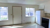 This single-floor space presents a straightforward and functional layout, with the entry and kitchen area occupying a shared open zone. The front door opens directly into this combined space, which features light-toned tile flooring that extends throughout the visible area, contributing to a clean and practical aesthetic.

A window positioned adjacent to the entry door allows natural light to filter in, offering a view of the surrounding exterior environment. The window is fitted with a standard blind, providing basic light and privacy control. Below the window sits a baseboard heater, offering an efficient heating solution integrated into the wall.

The kitchen area includes a full-size white refrigerator situated along the wall, with cabinetry visible in the upper corner, suggesting additional storage and workspace nearby. The walls are finished in a neutral off-white tone, and the ceiling features a textured finish. The overall interior palette is understated and utilitarian, emphasizing functionality. The space is compact in scale but makes efficient use of its layout, with all visible elements oriented toward practical everyday use.