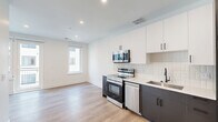 This thoughtfully designed unit offers a clean, contemporary aesthetic throughout its efficiently organized single-floor layout. The open kitchen serves as the centerpiece of the main living area, featuring a two-tone cabinet design with crisp white upper cabinets and dark lower cabinetry accented by matte black hardware. A white subway tile backsplash adds texture, while stainless steel appliances — including a range, over-the-range microwave, dishwasher, and refrigerator — provide a fully equipped cooking space. A single-basin undermount sink completes the kitchen. Light wood-tone flooring flows throughout, complementing the bright white walls and recessed lighting that keep the space feeling open and airy. A window in the living area allows natural light to filter in. The bathroom is a design highlight, featuring striking terrazzo-style flooring in a dark speckled pattern, vertical tile surrounding the soaking tub and shower, mustard-toned accent tiles above, and matte black fixtures that add a bold, modern touch. The private balcony opens to views of the surrounding multi-story building's courtyard, featuring metal railings and a spacious outdoor area. Additional conveniences include a dedicated closet and a utility room, rounding out this well-appointed space.