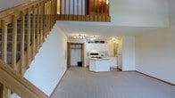 This two-story space showcases a loft-style design with an open-concept main floor that integrates the living and kitchen areas seamlessly. The living area features neutral carpeting and crisp white walls, creating a clean and airy atmosphere. A natural wood staircase with matching balustrade leads to the upper level, which overlooks the main floor through an open railing, adding vertical dimension and a sense of spaciousness to the interior.

The kitchen is outfitted with white cabinetry, a central island that provides additional workspace and counter space, and full-size appliances including a refrigerator and range. Light-toned flooring in the kitchen contrasts with the carpeted living area, helping to visually define each zone. Track lighting and flush-mount ceiling fixtures provide consistent illumination throughout the main floor.

A door near the base of the staircase leads to a storage closet, adding practical functionality to the layout. The upper loft level, visible from below, offers additional living space that benefits from the open sightlines to the main floor, enhancing the overall sense of connectivity throughout the home.