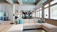 This single-floor space showcases a thoughtfully designed open-concept layout that seamlessly connects its living, dining, and lounge areas. The interior draws the eye immediately with its striking coffered ceiling detail, accented in a vibrant teal that complements the coastal-inspired color palette carried throughout the space. Crisp white structural columns define zones without interrupting the sense of openness, while large windows along the perimeter flood the interior with natural light and offer views of the surrounding outdoors. The lounge area is anchored by a generous sectional sofa in a neutral cream tone, dressed with layered throw pillows in shades of teal, gold, and blue, adding warmth and texture. Beyond the seating area, a dining space is elegantly appointed with deep navy blue furnishings and pendant lighting, creating a more intimate atmosphere within the broader open floor plan. The overall ambiance strikes a balance between relaxed and refined, with a cohesive design language that blends soft neutrals with bold accent tones. Recessed lighting throughout supplements the natural light, ensuring the space remains bright and welcoming at all hours. This is a well-appointed, inviting communal environment suited for both relaxation and social gatherings.