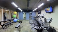 This fitness center is a dedicated single-floor workout facility designed to accommodate a variety of exercise needs. The space features an open layout with patterned carpeted flooring and exposed ceiling ductwork, lending it an industrial yet functional aesthetic. Bright fluorescent lighting illuminates the entire area, ensuring excellent visibility throughout.

The cardio zone is prominently featured along one wall and is lined with a row of modern elliptical machines and treadmills, each equipped with individual display screens. Wall-mounted televisions provide entertainment for users during their workouts. Full-length mirrors are strategically positioned along the blue-painted walls, enhancing the sense of space and allowing users to monitor their form.

The strength and functional training area includes an adjustable weight bench and a well-stocked rack of exercise balls in multiple sizes, foam rollers, and additional accessories, making it ideal for stretching, core work, and dynamic training. The combination of cardio equipment, strength tools, and functional fitness accessories ensures that this facility caters to a wide range of fitness levels and goals. The overall ambiance is energetic and practical, providing a focused environment well-suited for daily training.