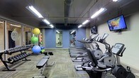 This fitness center is a dedicated single-floor workout facility designed to accommodate a variety of exercise needs. The space features an open layout with patterned carpeted flooring and exposed ceiling ductwork, lending it an industrial yet functional aesthetic. Bright fluorescent lighting illuminates the entire area, ensuring excellent visibility throughout.

The cardio zone is prominently featured along one wall and is lined with a row of modern elliptical machines and treadmills, each equipped with individual display screens. Wall-mounted televisions provide entertainment for users during their workouts. Full-length mirrors are strategically positioned along the blue-painted walls, enhancing the sense of space and allowing users to monitor their form.

The strength and functional training area includes an adjustable weight bench and a well-stocked rack of exercise balls in multiple sizes, foam rollers, and additional accessories, making it ideal for stretching, core work, and dynamic training. The combination of cardio equipment, strength tools, and functional fitness accessories ensures that this facility caters to a wide range of fitness levels and goals. The overall ambiance is energetic and practical, providing a focused environment well-suited for daily training.