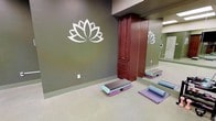 This tranquil wellness studio is thoughtfully designed to cultivate a calm and focused atmosphere for yoga and mindful movement. The space is defined by sage green walls that evoke a sense of serenity, anchored by a prominent lotus flower wall graphic — a universally recognized symbol of wellness and balance. A full-length mirrored wall runs along one side of the room, reflecting the space and allowing practitioners to monitor their form during sessions. The flooring is a smooth, neutral-toned surface that is both practical and easy to maintain, providing a clean foundation for exercise mats and equipment. A rich, dark wood storage cabinet with multiple drawers offers convenient in-room storage for supplies and accessories. The studio is equipped with exercise step platforms, yoga bolsters, and purple exercise mats, making it ready for a variety of fitness and wellness activities. Recessed lighting provides even, consistent illumination throughout the room, contributing to the peaceful ambiance. The overall design strikes a balance between functionality and mindful aesthetics, creating an inviting environment suitable for yoga classes, stretching, meditation, and low-impact fitness activities.