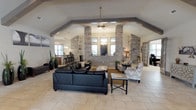 This single-story space is defined by its dramatic vaulted ceilings accented with prominent exposed wood beams, creating an open and airy atmosphere throughout. The main gathering area is generously sized, featuring large-format tile flooring and multiple distinct seating arrangements. A central lounge zone is anchored by a leather sofa and patterned accent chairs, while a second seating area near the entry showcases rich red leather furniture and large windows that allow natural light to fill the room.

Structural stone columns serve as a striking architectural focal point, dividing the main lounge from a bar and kitchenette area. This functional space features a granite-topped bar counter with barstool seating, pendant lighting, and a stone accent wall. Adjacent to the main lounge, a dining area with a solid wood pub-height table connects to a dedicated game room through oversized sliding barn doors. The game room contains a pool table and additional seating, making it well-suited for recreational use.

Ceiling fans are integrated throughout the open layout to complement the soaring roofline. The combination of stone, wood, and neutral tones gives the space a cohesive, rustic-contemporary character well-suited to communal gatherings.