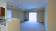 This single-floor space features a open living area finished with wall-to-wall carpeting in a neutral gray tone, providing a clean and practical foundation. The room is defined by crown molding along the ceiling line, adding a subtle architectural detail that enhances the overall finish of the space. Walls are painted in a warm beige tone, contributing to a bright and welcoming atmosphere. A sliding glass door dominates one wall of the living area, allowing natural light to fill the room and providing access to the outdoors. A floor-mounted heating vent is positioned beneath the sliding door, ensuring efficient climate control near the exterior wall. A partial view of an adjacent counter or bar area is visible to one side, suggesting a connected kitchen or dining space nearby. The ceiling features a recessed tray detail, adding dimension and visual interest overhead. Smoke detector and ventilation fixtures are integrated into the ceiling discreetly. The overall layout is straightforward and functional, offering an open, versatile living space with clean finishes throughout.