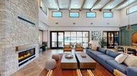 Located on Clinton Keith Road in Murrieta, California, this single-floor community space offers a thoughtfully designed interior that blends warmth, sophistication, and functionality. The living room serves as the heart of the space, showcasing soaring double-height ceilings with exposed wood beams, an impressive floor-to-ceiling stacked stone fireplace, and an abundance of natural light streaming through clerestory windows. Herringbone-patterned hardwood-style flooring adds visual depth throughout, complementing the neutral, earthy color palette. A striking arched entryway with decorative ironwork adds architectural character to the living area, which is furnished with a mix of navy sofas, leather armchairs, and natural wood accent tables. The dining room features a generous communal table beneath woven rattan pendant lights, with direct access to an outdoor area overlooking a resort-style pool — ideal for gatherings. A kitchenette with warm wood cabinetry, marble-look countertops, a matte black faucet, and a decorative arched tile alcove with open shelving provides functional support for events. Sage green millwork accents and high ceilings throughout give the space a refined yet inviting ambiance, making it a versatile and elegant setting for residents to gather and connect.