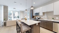 This single-floor residence showcases a thoughtfully designed open-concept layout with a contemporary aesthetic throughout. The kitchen serves as a focal point, featuring two-tone cabinetry with crisp white upper cabinets and warm wood-grain lower cabinets, a textured tile backsplash, and a full suite of stainless steel appliances including a refrigerator, electric range, built-in microwave, and dishwasher. A generously sized central island with a white quartz countertop provides ample prep space and casual seating for multiple guests, illuminated by a pair of pendant lights overhead. Light wood-style flooring runs throughout the main living areas, enhancing the bright, airy feel.

The bedrooms offer a calm, neutral palette with plush carpeting and large windows that invite natural light and outdoor views of the surrounding neighborhood. A exposed concrete accent wall in one bedroom adds an industrial design element. The bathroom is finished with a wide dual-sink vanity featuring wood-grain cabinetry, white quartz countertops, a frameless mirror, and light wood-style flooring, maintaining the cohesive modern style present throughout the entire residence.
