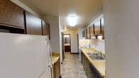 Located at 1500 S Adams Ave in Marshfield, Wisconsin, this single-floor unit offers a straightforward, functional layout well-suited for everyday living. The galley-style kitchen runs along a narrow corridor, lined on both sides with dark wood cabinetry providing ample storage. Light wood laminate countertops span the length of the kitchen, complemented by a double-basin stainless steel sink, an electric range, and a dishwasher. Durable tile-style flooring completes the kitchen area, and a hallway from the kitchen leads directly to the bathroom.

The bathroom includes a vanity with a sink, a toilet, an over-toilet storage cabinet, and a tub-shower combination enclosed by a curtain. The living areas and bedrooms feature wall-to-wall carpeting, creating a comfortable feel underfoot. Ceiling fans with integrated light fixtures are present in the living room and entry area, providing both air circulation and ambient lighting. Baseboard heating units run along the walls throughout the carpeted rooms, ensuring warmth during Wisconsin winters. Bedrooms include windows dressed with vertical blinds, allowing natural light while maintaining privacy. The front entry opens directly into the main living area, which offers a generous, open feel suitable for a variety of furniture arrangements.