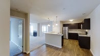This single-floor residence is designed with an open-concept layout that promotes a sense of spaciousness and connectivity between living areas. The kitchen is a standout feature, showcasing rich dark cabinetry that contrasts elegantly with light granite countertops. A central island with an integrated sink serves as both a functional workspace and a casual gathering spot, illuminated by a trio of stylish pendant lights that hang overhead. Stainless steel appliances, including a full-size refrigerator, add a sleek, modern touch. The kitchen flows naturally into the living area, where warm hardwood-style flooring transitions to plush wall-to-wall carpeting. The living room is bright and inviting, benefiting from natural light streaming through large windows and a set of glass panel doors. A fireplace serves as a focal point, adding warmth and character to the space. The neutral color palette throughout — soft whites, warm beiges, and deep espresso tones — creates a cohesive and welcoming ambiance. The textured ceilings and clean white trim add subtle architectural detail, completing the well-appointed feel of this thoughtfully designed single-level home.