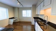 This single-floor space offers a functional and practical layout designed for comfortable everyday living. The living area is finished with neutral-toned carpet and light beige walls, complemented by warm wood baseboard trim. A wall-mounted heating and ventilation unit provides climate control, while a window fitted with blinds allows natural light to fill the room. A hallway connects the living area to the rest of the unit, offering a logical and easy-to-navigate floor plan.

The kitchen is a standout feature, showcasing white cabinetry paired with striking dark granite-style countertops that provide a bold contrast. A stainless steel sink and dishwasher add to the kitchen's functionality. A dedicated built-in desk area with matching dark countertop surface sits adjacent to the kitchen, making it an ideal spot for a home office or study nook. Hardwood-style flooring runs throughout the kitchen and dining area, adding warmth and visual interest. Track lighting mounted on the textured ceiling provides targeted illumination across the space.

Windows with blinds in both the living and kitchen areas invite natural light and give the space an open, airy feel. Throughout, the neutral color palette and clean finishes create a versatile environment ready for personalization.