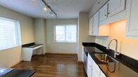 This single-floor space offers a functional and practical layout designed for comfortable everyday living. The living area is finished with neutral-toned carpet and light beige walls, complemented by warm wood baseboard trim. A wall-mounted heating and ventilation unit provides climate control, while a window fitted with blinds allows natural light to fill the room. A hallway connects the living area to the rest of the unit, offering a logical and easy-to-navigate floor plan.

The kitchen is a standout feature, showcasing white cabinetry paired with striking dark granite-style countertops that provide a bold contrast. A stainless steel sink and dishwasher add to the kitchen's functionality. A dedicated built-in desk area with matching dark countertop surface sits adjacent to the kitchen, making it an ideal spot for a home office or study nook. Hardwood-style flooring runs throughout the kitchen and dining area, adding warmth and visual interest. Track lighting mounted on the textured ceiling provides targeted illumination across the space.

Windows with blinds in both the living and kitchen areas invite natural light and give the space an open, airy feel. Throughout, the neutral color palette and clean finishes create a versatile environment ready for personalization.