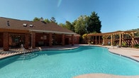 This community amenity space centers around a generously sized freeform swimming pool with clear, blue water and a smooth surrounding deck ideal for lounging. The pool is flanked by a classic brick clubhouse with a covered porch supported by brick columns, offering shaded seating and a welcoming outdoor retreat. A wooden pergola on the opposite side of the pool adds a charming architectural accent and additional outdoor gathering space, all set against a backdrop of mature trees under open skies.

The interior of the clubhouse is equally well-designed, featuring soaring vaulted ceilings with crown molding and recessed lighting that create an open, airy atmosphere. Rich hardwood flooring runs throughout, complementing the neutral gray walls and white trim. The main lounge area is furnished with plush tufted gray armchairs arranged around accent tables, providing a comfortable setting for socializing or relaxing. Artwork adorns the walls, adding a polished, curated feel to the space.

A built-in bar-height workstation with computer monitors serves as a convenient resident services area. Adjacent to the lounge, a pass-through kitchen features white cabinetry, black appliances, a breakfast bar with wire barstools, and a ceiling-mounted pendant light. A secondary room off the kitchen provides additional utility space, completing this thoughtfully equipped community facility.