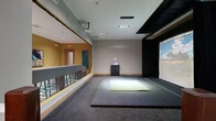 Located at 824 W Route 66 in Flagstaff, Arizona, this indoor golf simulator space offers a dedicated, self-contained bay designed for an immersive golf experience. The main simulator area is carpeted in dark grey and centers around a large projection screen mounted against a black-draped wall, displaying realistic golf course visuals. A bright artificial turf hitting mat is positioned on the floor directly in front of the screen, providing the designated swing area. An overhead projector is ceiling-mounted to deliver sharp, full-screen imagery, while a compact control console cabinet sits nearby for managing simulator settings. Recessed ceiling lighting illuminates the space evenly throughout. Adjacent to the simulator bay is a separate viewing and waiting corridor, finished with a distinctive checkered tile floor in muted tones and warm wood base trim. This side area is lined with a low partition wall featuring wood-cap railing, allowing observers a clear sightline into the simulator. The overall layout is clean and functional, with neutral grey walls, wood accents, and clear delineation between the active hitting zone and the spectator corridor. The space is well-suited for individual practice sessions or small group play in a controlled, year-round indoor environment.