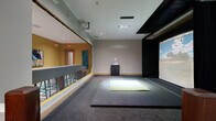 Located at 824 W Route 66 in Flagstaff, Arizona, this indoor golf simulator space offers a dedicated, self-contained bay designed for an immersive golf experience. The main simulator area is carpeted in dark grey and centers around a large projection screen mounted against a black-draped wall, displaying realistic golf course visuals. A bright artificial turf hitting mat is positioned on the floor directly in front of the screen, providing the designated swing area. An overhead projector is ceiling-mounted to deliver sharp, full-screen imagery, while a compact control console cabinet sits nearby for managing simulator settings. Recessed ceiling lighting illuminates the space evenly throughout. Adjacent to the simulator bay is a separate viewing and waiting corridor, finished with a distinctive checkered tile floor in muted tones and warm wood base trim. This side area is lined with a low partition wall featuring wood-cap railing, allowing observers a clear sightline into the simulator. The overall layout is clean and functional, with neutral grey walls, wood accents, and clear delineation between the active hitting zone and the spectator corridor. The space is well-suited for individual practice sessions or small group play in a controlled, year-round indoor environment.