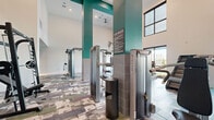 Located at 7222 S Peek Rd in Houston, TX, this expansive fitness center is a well-appointed, single-floor facility designed to support a full range of workout routines. The space makes an immediate impression with its soaring double-height ceilings accented by bold teal structural columns, creating an open and energizing atmosphere throughout.

The main floor is generously proportioned, accommodating a diverse mix of Matrix-brand strength training machines, free weight stations, a Smith machine, and a row of cardio equipment including treadmills and a Jacob's Ladder stair climber. A mezzanine level, accessible via an open-tread wooden staircase with sleek cable railings, provides additional workout space and overlooks the main floor below.

Large windows along the perimeter flood the interior with natural light and offer views of the surrounding landscape, enhancing the sense of openness. The floors are covered in a vibrant, multi-toned patchwork-patterned carpet tile that adds a dynamic visual element to the space.

An artistic accent wall featuring a colorful, abstract bubble-motif wallcovering adds a creative focal point near the staircase. The overall design balances functionality with contemporary aesthetics, making this fitness center an inviting and well-equipped destination for residents and users in the Richmond/Houston area.