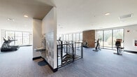 This fitness center offers a thoughtfully designed, expansive single-floor layout that accommodates a comprehensive range of exercise equipment and dedicated workout zones. The main cardio area features rows of treadmills and stationary bikes positioned along a wall of large floor-to-ceiling windows, flooding the space with natural light and providing open views of the surrounding landscape. A prominent exposed brick column serves as a distinctive architectural accent, adding warmth and character against the otherwise clean, modern aesthetic of white walls and recessed lighting.

A separate cycling studio is enclosed by sleek glass-panel partitions with dark steel frames, creating a private yet visually connected workout zone. This room features spin bikes, a large wall mirror that enhances the sense of space, medicine balls, stability balls, and a tray ceiling with ambient accent lighting that lends an upscale atmosphere.

Throughout the fitness center, durable blue-toned carpet flooring provides comfort underfoot, while an interior corridor connects the various zones with ease. A water fountain is conveniently located near the stairwell entry point, which features contemporary metal railings. The overall design balances functionality with modern aesthetics, creating an inviting and well-lit environment suited for a full fitness regimen.