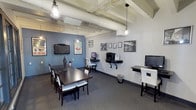 This single-floor multipurpose room blends industrial design with functional amenities, creating a practical and visually engaging environment. The space features an exposed ceiling with visible ductwork and structural beams painted white, complemented by pendant cage-style light fixtures that lend an urban, loft-like character. The flooring is covered in dark gray carpet, adding a grounded, professional feel throughout.

A central dark wood conference table with white upholstered chairs serves as the primary gathering area, suitable for meetings or collaborative work. A wall-mounted flat-screen television is positioned above on a blue accent wall, which also features a decorative Monaco racing poster, adding a touch of personality to the space.

Along the adjacent light gray wall, wall-mounted shelving supports computer workstations, a printer, and additional monitors, providing practical business center functionality. A series of framed black-and-white photographs line the walls, contributing to the space's curated aesthetic. The overall ambiance is clean, organized, and well-equipped, balancing both social and productivity-oriented uses effectively.