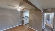 This single-floor space offers a practical and straightforward layout designed for comfortable everyday living. The open-concept design seamlessly connects the main living area to the kitchen, creating a sense of spaciousness and ease of movement throughout. The flooring is finished in warm hardwood-style planks that extend across the main living space, adding a clean and cohesive look underfoot. The kitchen is compact yet functional, featuring white cabinetry, a single-basin sink, and ample counter space for meal preparation. The white cabinetry provides a bright contrast against the neutral grey walls, keeping the kitchen area feeling light and tidy. A structural beam detail adds a subtle architectural element to the ceiling, giving the space a bit of visual character. The living area is equipped with a ceiling fan with an integrated light fixture, ensuring comfort and adequate lighting throughout the day and evening. Textured ceilings are present throughout, and the neutral grey wall tones provide a versatile backdrop suited to a variety of décor styles. Overhead ventilation is also in place, supporting air circulation and climate control. A doorway visible from the main area suggests additional rooms or storage spaces accessible from the central living zone.