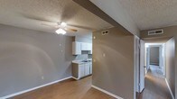 This single-floor space offers a practical and straightforward layout designed for comfortable everyday living. The open-concept design seamlessly connects the main living area to the kitchen, creating a sense of spaciousness and ease of movement throughout. The flooring is finished in warm hardwood-style planks that extend across the main living space, adding a clean and cohesive look underfoot. The kitchen is compact yet functional, featuring white cabinetry, a single-basin sink, and ample counter space for meal preparation. The white cabinetry provides a bright contrast against the neutral grey walls, keeping the kitchen area feeling light and tidy. A structural beam detail adds a subtle architectural element to the ceiling, giving the space a bit of visual character. The living area is equipped with a ceiling fan with an integrated light fixture, ensuring comfort and adequate lighting throughout the day and evening. Textured ceilings are present throughout, and the neutral grey wall tones provide a versatile backdrop suited to a variety of décor styles. Overhead ventilation is also in place, supporting air circulation and climate control. A doorway visible from the main area suggests additional rooms or storage spaces accessible from the central living zone.