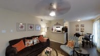 This single-story space is designed with an open-concept layout that seamlessly connects the living area, dining space, and kitchen. The living room serves as the central hub, featuring warm beige walls, dark hardwood-style flooring, and a ceiling fan with integrated lighting that enhances the room's comfortable ambiance. Tasteful wall art adorns the walls, adding a decorative touch to the neutral palette.

The kitchen is accessible via a raised breakfast bar counter, which doubles as a casual dining or prep surface. Dark appliances, including a refrigerator and range, are visible from the living area, and white cabinetry provides a clean contrast. A hallway beyond the kitchen suggests additional rooms within the unit.

The living area is furnished with a dark upholstered sofa accented with orange and floral throw pillows, complemented by a tufted blue accent chair that adds visual interest. A glass coffee table and a floor lamp contribute to the inviting atmosphere. A separate dining area is partially visible to the right, completing this well-appointed, cohesive living space.