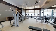 This fitness center offers a thoughtfully designed workout environment that balances functionality with contemporary style. The space is anchored by large, floor-to-ceiling windows along one wall, flooding the interior with natural light and providing open views to the outside. A raised ceiling section near the windows enhances the sense of vertical space, while recessed lighting and dual ceiling fans ensure the room remains well-lit and comfortable during workouts. The interior design incorporates a striking contrast between dark wall accents and light-toned flooring, creating a polished, modern aesthetic. A decorative mural featuring vintage athletic imagery adds character to one wall. The fitness floor is well-organized, housing a comprehensive selection of equipment including cable and weight machines, elliptical trainers, treadmills, and a weight bench, catering to a range of strength and cardiovascular training needs. The light-colored flooring is durable and easy to maintain, contributing to the overall clean appearance of the space. The layout allows for comfortable movement between stations, making the facility practical for individual workouts.
