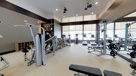 This fitness center offers a thoughtfully designed workout environment that balances functionality with contemporary style. The space is anchored by large, floor-to-ceiling windows along one wall, flooding the interior with natural light and providing open views to the outside. A raised ceiling section near the windows enhances the sense of vertical space, while recessed lighting and dual ceiling fans ensure the room remains well-lit and comfortable during workouts. The interior design incorporates a striking contrast between dark wall accents and light-toned flooring, creating a polished, modern aesthetic. A decorative mural featuring vintage athletic imagery adds character to one wall. The fitness floor is well-organized, housing a comprehensive selection of equipment including cable and weight machines, elliptical trainers, treadmills, and a weight bench, catering to a range of strength and cardiovascular training needs. The light-colored flooring is durable and easy to maintain, contributing to the overall clean appearance of the space. The layout allows for comfortable movement between stations, making the facility practical for individual workouts.