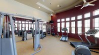 This fitness center is designed to accommodate a wide range of workout preferences within a spacious, single-floor layout. The architecture is immediately striking, with soaring tray ceilings, recessed lighting, and dual ceiling fans that keep the space comfortable and well-ventilated. Bold crimson accent walls contrast with warm neutral tones throughout, creating an energizing yet polished atmosphere.

A curved bank of floor-to-ceiling windows along one wall floods the interior with natural light and offers views of an outdoor pool area, making cardio sessions particularly enjoyable. High clerestory windows on additional walls further enhance the bright, open feel of the space.

The cardio zone is well-appointed with multiple Precor elliptical machines, treadmills, and a stair stepper, all positioned to take advantage of the natural light. A mounted flat-screen television provides entertainment during workouts. The strength-training area includes a multi-station cable machine, a dedicated free weight section with tiered dumbbell racks, adjustable benches, and a punching bag for functional training. Large floor-to-ceiling mirrors visually expand the weight area while allowing users to monitor their form. The flooring transitions thoughtfully between carpet and hard surfaces based on the functional demands of each zone.