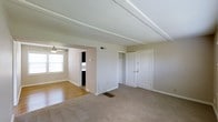 This single-floor residence offers a straightforward and functional layout designed for comfortable everyday living. The interior is finished with a neutral color palette of soft whites and warm beiges, creating a bright and welcoming atmosphere throughout.

The main living area is generously sized, finished with wall-to-wall carpeting that adds warmth underfoot. Exposed beam detailing along the ceiling lends a subtle architectural character to the space. A floor vent indicates central climate control, ensuring year-round comfort.

An open passageway connects the carpeted living area to an adjoining room featuring hardwood flooring, which brings a contrasting warmth and texture to the space. This secondary area is brightened by a window that allows natural light to filter in, complemented by a ceiling-mounted light fixture. The transition between the two areas is defined by a partial wall, adding a sense of separation while maintaining an open, connected feel.

A set of double doors leads to a built-in closet, providing ample storage space, while an additional door offers access to a separate room or utility area. Clean white trim and baseboards run consistently throughout, tying the rooms together cohesively.

Overall, the space presents a clean, move-in-ready interior with practical storage solutions and a calm, neutral ambiance.