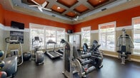 Located in Menifee, CA, this fitness center is a thoughtfully designed exercise space that combines energetic aesthetics with a comprehensive range of workout equipment. The room immediately draws the eye with its bold orange upper walls, contrasted by a neutral lower tone, creating a vibrant and motivating atmosphere. A striking coffered ceiling adds architectural character overhead, while a ceiling fan ensures comfortable airflow throughout workouts.

Natural light pours in generously through multiple large windows fitted with blinds, offering views of the outdoor surroundings and keeping the space feeling open and inviting. Full-length mirrors along one wall visually expand the room and provide functional feedback during strength training sessions.

The exercise room is well-stocked with a diverse selection of equipment. Cardio enthusiasts will find treadmills, elliptical trainers, and a stationary bike ready for use. The strength training area features a multi-functional cable machine, a dedicated weight bench, a comprehensive dumbbell rack with a wide range of free weights, and a plate-loaded rack. Medicine balls are also available for functional fitness routines.

A wall-mounted television is positioned for convenient viewing during cardio sessions, and rubber flooring throughout provides durability, safety, and comfort underfoot. The space is well-lit with recessed lighting complementing the natural light, making it a fully functional and welcoming facility for a complete workout experience.