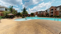 This expansive outdoor amenity space is designed to deliver a resort-like experience for its residents. At the heart of the area is a generously sized freeform swimming pool with clear turquoise water and a decorative central fountain, creating a tranquil and visually striking focal point. The pool is framed by a broad stamped concrete sundeck that provides ample space for lounge chairs and open-air relaxation under the open sky.

A substantial wood-beam pergola anchors the outdoor living area, offering shaded zones equipped with bar-height dining tables and chairs — ideal for social gatherings and al fresco meals. Adjacent to the pergola, a dedicated outdoor kitchen features dual built-in stainless steel grills mounted on a brick base, providing a convenient and functional space for barbecuing and entertaining.

The surrounding landscape is thoughtfully maintained, with tropical palm trees, flowering crepe myrtles in vibrant pink, manicured hedges, and lush ground cover adding color and a warm, welcoming atmosphere. The multi-story brick residential buildings that frame the courtyard lend a cohesive architectural character to the space. Overall, the amenity area combines practical outdoor living features with attractive landscaping to create an inviting communal environment.