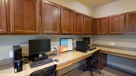 This single-floor business center is thoughtfully designed to provide a functional and efficient workspace. The room features a continuous built-in countertop workstation that wraps along the walls, offering generous desk space for multiple users simultaneously. Each workstation is equipped with a desktop computer setup, accommodating the needs of those requiring reliable access to technology and computing resources.

Overhead, a full run of rich, dark wood cabinetry with raised-panel doors extends across the length of the workspace, providing substantial closed storage that keeps the environment neat and clutter-free. The warm tones of the cabinetry contrast with the light-colored countertop surface, lending the room a professional yet inviting atmosphere.

Ergonomic task chairs are positioned at each workstation, ensuring comfort during extended use. The flooring is finished in a neutral carpet, contributing to the room's quiet and focused ambiance. The overall layout is practical and space-conscious, making efficient use of every available square foot. Whether used for business correspondence, research, or general computing needs, this business center offers a well-appointed and orderly environment suited to a variety of professional tasks.
