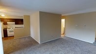 This single-floor space features an open, functional layout with a neutral color palette that creates a clean, understated atmosphere. The main living area is finished with wall-to-wall carpeting in a light grey tone, providing a soft and comfortable underfoot experience. Walls are painted in warm beige and tan hues, contributing to a cohesive and inviting interior aesthetic.

A defining architectural element of the space is a centrally positioned partial wall that divides the interior without fully enclosing it, maintaining a sense of openness while subtly delineating separate zones within the floor plan. This structural feature adds visual interest and offers practical spatial organization.

To one side, a glimpse of the kitchen area is visible, where darker cabinetry contrasts with the lighter wall tones. A doorway on the opposite side leads to additional areas of the unit, suggesting further rooms beyond the main living space. White baseboards trim the perimeter of the room, adding a crisp finish to the overall design. The standard ceiling height and straightforward layout make this a practical and adaptable space suited to a variety of furnishing arrangements.